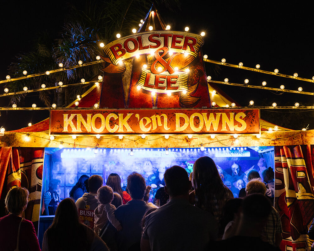 A crowd waiting in line to walk into a brightly lit miniature circus tent. A Red and yellow sign says "Knock 'em Downs".